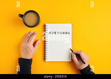 Hände eines Mannes bereit, in seine Notizblock mit dem Wort Estate Planung mit einer Tasse Tee neben ihm zu schreiben. Gelber Hintergrund über dem Kopf. Stockfoto