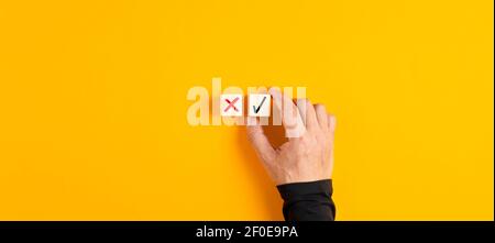 Richtige und falsche Symbole auf Holzwürfeln mit männlicher Hand die Wahl der richtigen Symbol auf gelbem Hintergrund. Genehmigen, Abstimmen oder richtiges Entscheidungskonzept. Stockfoto