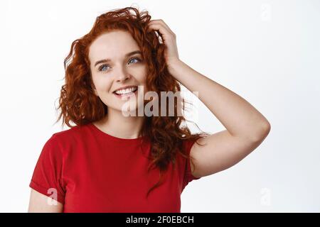 Portrait von verträumten niedlichen Teenager-Mädchen mit roten lockigen Haaren, lächelnd und Blick auf die linke Kopie Raum, berühren gesunde natürliche Ingwer Haarschnitt, weiß Stockfoto