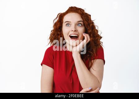 Porträt einer faszinierten jungen Frau mit rotem Lockengehirn, keuchend erstaunt, starrend aufgeregt auf der linken Seite mit Kopieplatz für Ihr Logo oder Banner, stehend Stockfoto