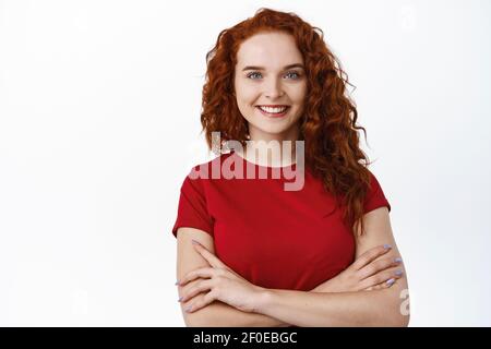 Ich bin zuverlässig. Selbstbewusste junge Frau mit lockigen roten Haaren und guter Hautzustand, natürliche weiße Lächeln, Kreuzarme auf der Brust und Blick entschlossen Stockfoto