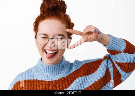 Nahaufnahme Porträt von glücklich und positiv Rotschopf Mädchen in der Brille, zeigt Zunge mit V-Zeichen und Spaß haben, Freude und Aufregung zum Ausdruck bringen, weiß Stockfoto