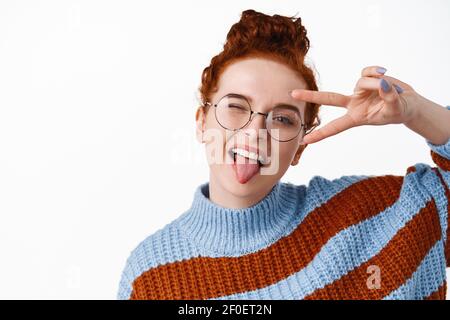 Nahaufnahme von positiven Hipster Mädchen mit roten Haaren gekämmt in Brötchen, Brille tragen, zeigt V-Zeichen Frieden und Augenzwinkern bei der Kamera mit erweiterter Zunge, weiß Stockfoto