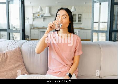 Attraktive asiatische Frau trinkt Wasser aus Glas sitzt auf dem Sofa zu Hause, stellt den Wasserhaushalt, Hydratation für Frische und gutes Aussehen. Routinevormittag Stockfoto