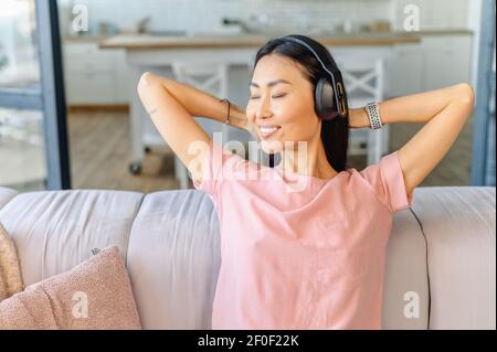 Eine zufriedene und fröhliche junge asiatische Frau mit Kopfhörern sitzt auf der Couch, ruht zu Hause und wirft ihre Hände hinter den Kopf, hört Musik und genießt das Wochenende Stockfoto