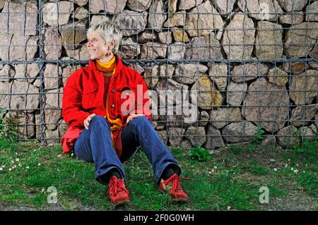 Ganzkörperportrait einer reifen Frau in blauer Jeans, roter Jacke, roten Sportschuhen und gelbem Schal sitzen auf dem Boden mit dem Rücken gegen einen verbarrt Stockfoto