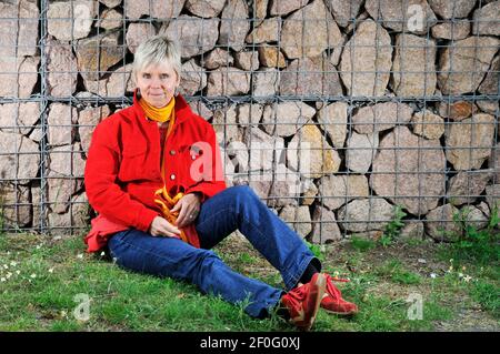 Ganzkörperportrait einer reifen Frau in blauer Jeans, roter Jacke, roten Sportschuhen und gelbem Schal sitzen auf dem Boden mit dem Rücken gegen einen verbarrt Stockfoto