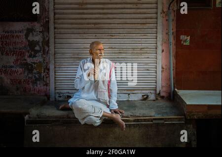 Aufsehenwürdiges Porträt eines älteren Mannes, der mit gekreuzten Beinen nachdenklich sitzt und in der Straßenszene in Mathura, Uttar Pradesh, Indien, Tee trinkt. Stockfoto