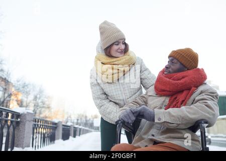 Porträt eines afroamerikanischen Mannes, der im Winter im Rollstuhl im Freien sitzt, mit lächelnder junger Frau, die ihn ansieht und ihm hilft, Raum zu kopieren Stockfoto