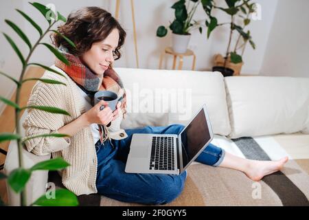 Komfortable schöne junge Frau beobachten einen Laptop in ihrer Wohnung. Sie sitzt auf einer Couch, trägt einen Schal und trinkt einen Kaffee. Seitenansicht. Stockfoto