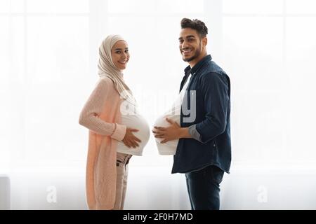 Junge schwangere muslimische Frau, die Spaß mit Ehemann zu Hause, Bäuche vergleichen Stockfoto