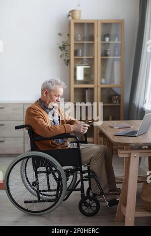 Vertikale Seitenansicht Porträt eines älteren Mannes im Rollstuhl mit Smartphone beim Sitzen am Schreibtisch in der Inneneinrichtung Stockfoto