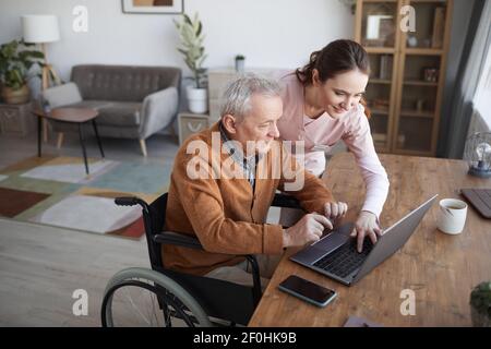 Porträt eines älteren Mannes im Rollstuhl mit Laptop im Altersheim mit Krankenschwester, die ihn unterstützt, kopieren Raum Stockfoto