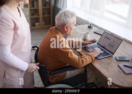 Hochwinkelporträt eines älteren Mannes im Rollstuhl mit Laptop im Altersheim mit Krankenschwester, die ihm hilft, Kopierraum Stockfoto