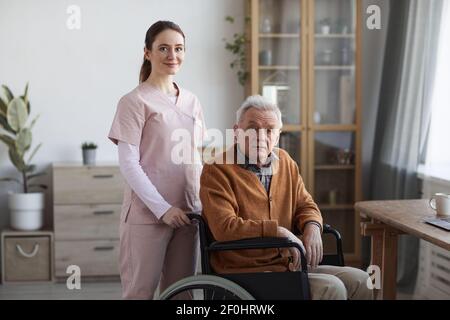 Porträt einer jungen Krankenschwester, die auf die Kamera schaut, während sie einem älteren Mann im Rollstuhl hilft, Raum kopieren Stockfoto