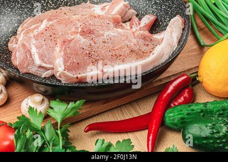 Stücke von frischem rohem Fleisch in einer dunklen Pfanne mit Gewürzen auf dem Hintergrund eines Schneidebretts neben Pilzen, Petersilie, Dill, Tomaten, grünen Zwiebeln, lemo Stockfoto
