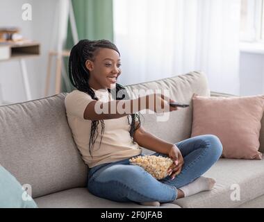 Glückliche junge schwarze Frau sitzt auf der Couch mit TV-Fernbedienung Stockfoto