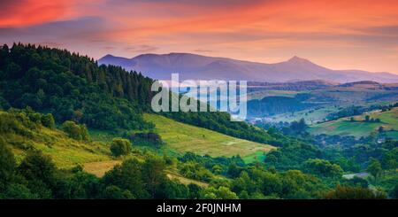 Bergige ländliche Landschaft bei Morgendämmerung. Schöne Landschaft mit Wäldern, Hügeln und Wiesen im Morgenlicht. Kamm mit hohen Gipfel in der Ferne. Dorf Stockfoto