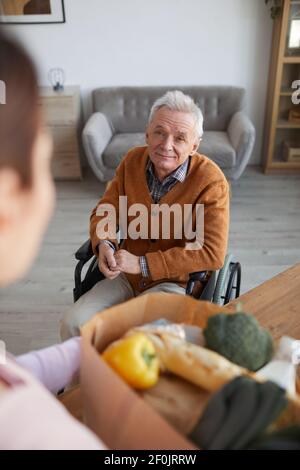 Hochwinkelporträt eines lächelnden älteren Mannes im Rollstuhl, der eine Krankenschwester ansieht, die Lebensmittel, Hilfe und ein Konzept für die Lieferung von Lebensmitteln bringt Stockfoto