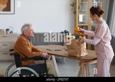 Seitenansicht Porträt eines lächelnden älteren Mannes im Rollstuhl, der eine Krankenschwester ansieht, die Lebensmittel, Hilfe und ein Konzept für die Lieferung von Lebensmitteln bringt Stockfoto