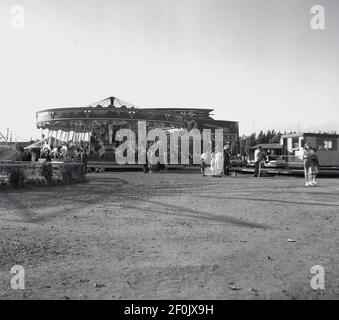 1950s, historisch, Blick über einen geschotterten Außenbereich, von einem Merry-Go-Round oder Karussell Spaß fair Ride, England, Großbritannien. Ein Vergnügungsritt für Kinder und Eltern, es ist eine drehbare kreisförmige Plattform mit Sitzplätzen für Reiter, traditionell in Form von Reihen von Holzpferden oder anderen Tieren auf Pfosten montiert, die auf und ab bewegen. Stockfoto