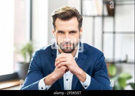 Erfolgreicher maskuliner Geschäftsmann in formell teuren Anzug, mit ordentlichen Stoppeln, sitzen, Hände vor ihm halten, Millennial selbstbewussten Mann posiert für Foto, Blick auf die Kamera, im Büro Hintergrund. Stockfoto