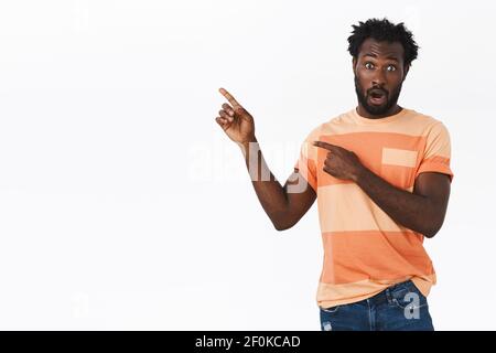 Überrascht und beeindruckt afroamerikanischen bärtigen Kerl in gestreiftem T-Shirt, offenen Mund fasziniert, keuchend schockiert, als zeigen obere linke Ecke starren Stockfoto