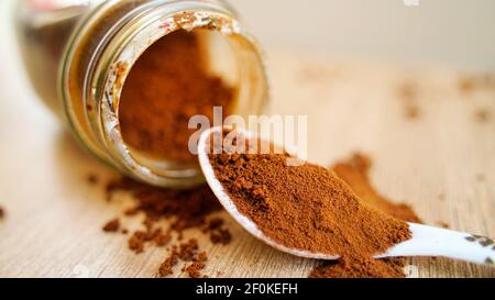 Kaffeepulver verschüttet auf dem Holztisch mit weißem Löffel. Stapel von braunem Kaffee mit einem Glas in unscharfen Hintergrund. Makroansicht von oben, Indien Stockfoto