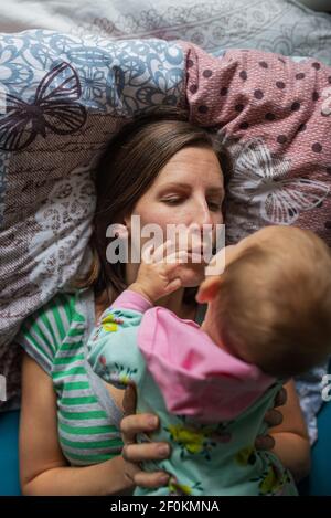 Die junge Mutter liegt auf dem Bett und ihre kleine Tochter liegt auf ihr in einem zarten Moment zwischen Mutter und Tochter. Stockfoto