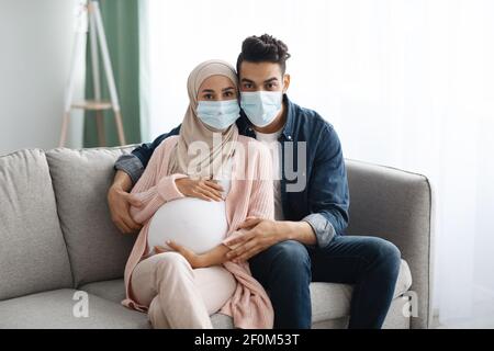 Junge schwangere muslimische Paar trägt medizinische Masken auf der Couch sitzen Zu Hause Stockfoto