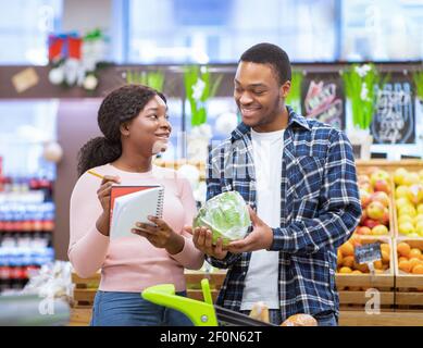Einkaufen während des Coronavirus-Ausbruchs. Millennial schwarzes Paar Kauf frisches Gemüse, Überprüfung der Lebensmittelliste im Einkaufszentrum Stockfoto