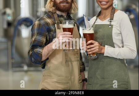 Beschnittenes Porträt von zwei lächelnden jungen Arbeitern, die Bierglas halten und die Kamera betrachten, während sie in der Werkstatt in der Brauerei stehen, Kopierraum Stockfoto