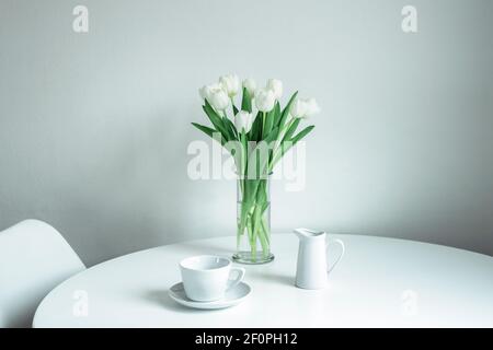 Bouquet von weißen Tulpen in einer Glasvase, einer Tasse und einem Milchkännchen auf einem weißen Tisch. Morgen, Frühstück, Frühlingskonzept. Stockfoto