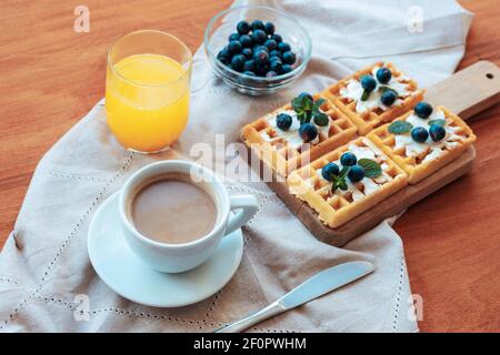 Waffeln mit Sahne und Heidelbeeren, eine Tasse Kaffee und Orangensaft auf dem Tisch auf einer Stoffserviette. Draufsicht. Stockfoto