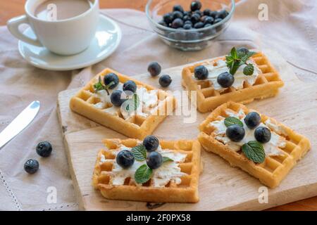 Waffeln mit Sahne und Heidelbeeren auf Schneidebrett, eine Tasse Kaffee. Süßes Essen, Frühstück. Draufsicht. Stockfoto