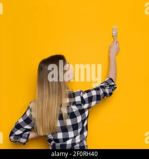 Junge blonde Frau im Holzfällerhemd ist Malerei gelbe Wand. Rückansicht. Hüfthöhe. Stockfoto