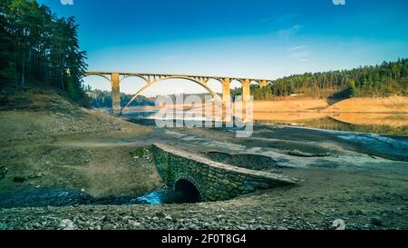 Hoch gewölbter Viadukt über leeren Orlik Damm und original Moldau Flussbett. Kontrast von großem Beton und kleiner alter Steinstraßebrücke. Podolsko, Tschechien. Stockfoto