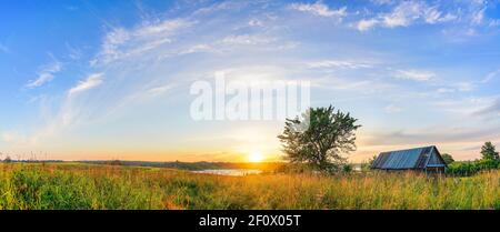 Panoramablick auf Sonnenuntergang über alte Holzhütte und einsam Baum auf dem Land Stockfoto