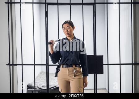 Asiatische Garde in Uniform in voller Rüstung mit einem Schlagstock Und eine Pistole auf dem Hintergrund einer Gefängniszelle Stockfoto