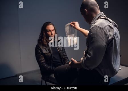 Im Verhörraum legt ein Beamter des Drogenkontrolldienstes dem Verbrecher Beweise für weißes Kokainpulver in einem Paket vor. Stockfoto