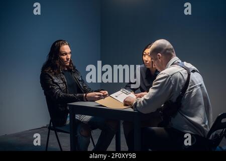 Ein zäher Polizeidetektiv verhört einen Tatverdächtigen mit einem tätowierten Gesicht in einem Verhörraum. Stockfoto