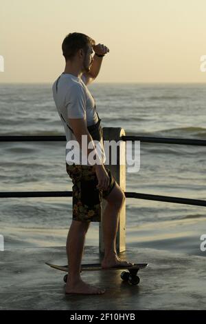 Junger erwachsener Mann, Skateboarder, aktiver Lebensstil, auf Pier stehend, Blick auf das Meer, Durban, Südafrika, Freizeitaktivitäten, Freizeitkleidung, sorglos Stockfoto