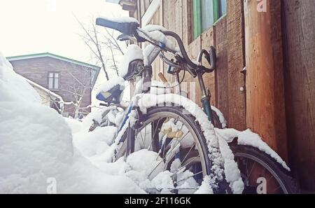 Mit Schnee bedeckte Fahrräder stehen an einer Holzwand in der Nähe des Hauses. Alte verlassene Fahrräder im Hinterhof eines Hauses unter dem Schnee Stockfoto