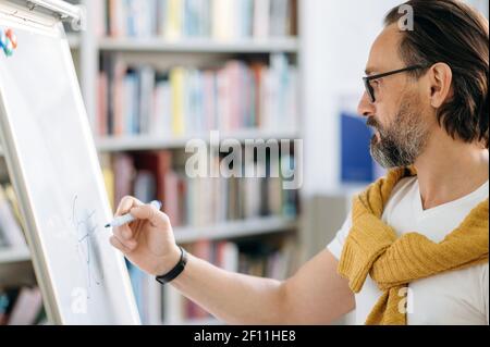 Konzentrierter kluger Kaukasier mittleren Alters, Lehrer oder Geschäftsleiter, schreibt Informationen auf einem Markiertafel während eines Vortrags Stockfoto