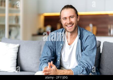 Porträt eines attraktiven charismatischen kaukasischen blauäugigen Kerl in einem Jeanshemd sitzt auf einem Sofa im Wohnzimmer Mit einem freundlichen Lächeln auf die Kamera schauen Stockfoto