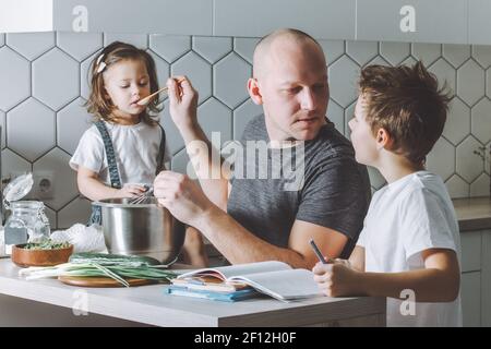 Vater peitscht Omelette mit Schneebesen, hilft seinem Sohn seine Hausaufgaben zu machen und füttert seine Tochter in der Küche. Mann, der Aufgaben erledigt. Stockfoto