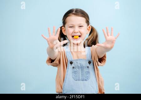 Lustige kleine Mädchen posiert mit Süßigkeiten in ihren Zähnen über blauen Hintergrund. Sie hat zwei Pferdeschwänze, trägt Overalls. Ihre Hände streckten sich mit dem Fortschreiten aus Stockfoto