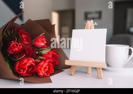 Ein Bouquet aus roten Tulpen auf dem Tisch und eine weiße Tasse Kaffee, leerer weißer Rahmen. Konzept für eine Grußkarte. Hochwertige Fotos Stockfoto