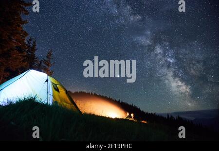 Zwei Menschen sitzen neben Lagerfeuer auf ihrem Campingplatz mit beleuchteten Zelt und schauen auf Flamme. Nacht Bergwald und Sternenhimmel auf dem Hintergrund. Stockfoto