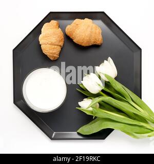Flaches Tablett mit zwei Croissants, einer Tasse Kaffee und weißen Tulpenblüten auf Weiß Stockfoto